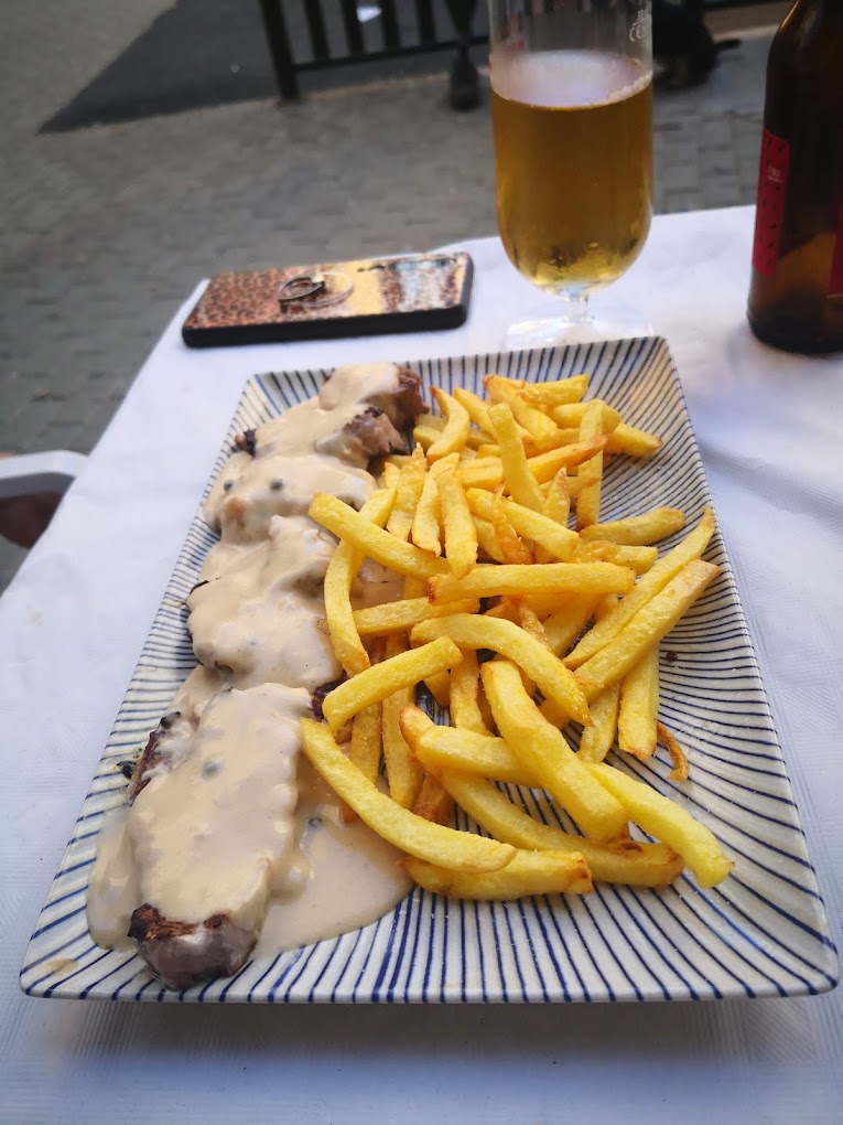 Plato de solomillo con salsa y patatas fritas junto a una cerveza en una mesa de terraza.