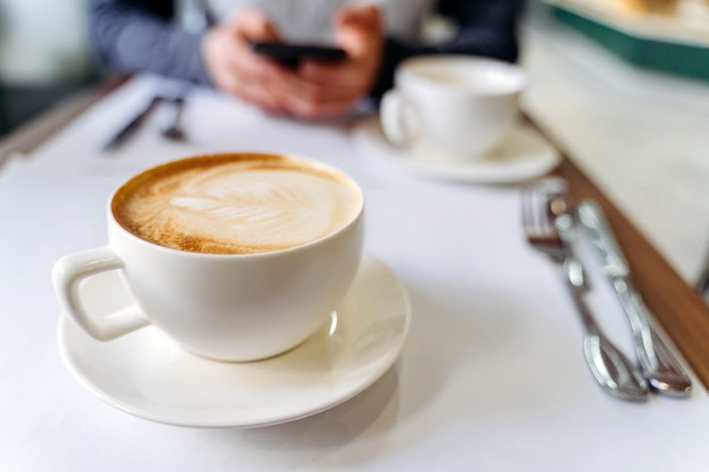 Taza de café con leche y arte latte sobre una mesa con una persona usando un teléfono al fondo.