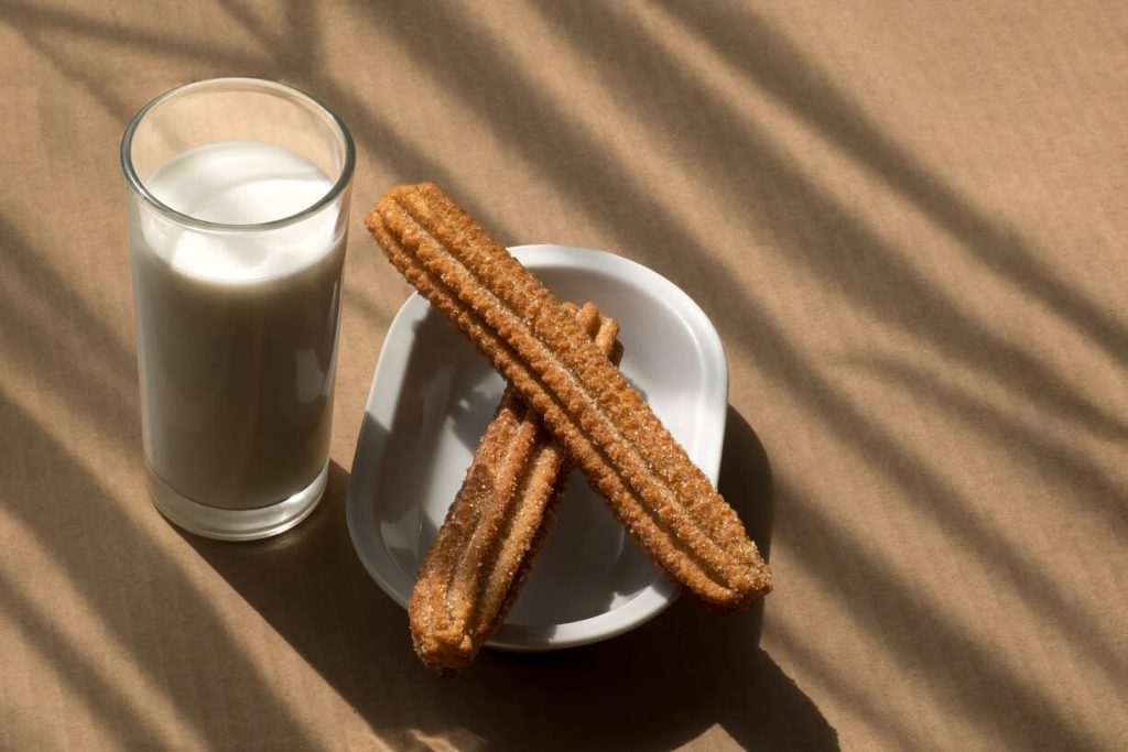 Churros recién hechos junto a un vaso de leche en una mesa con luz natural.