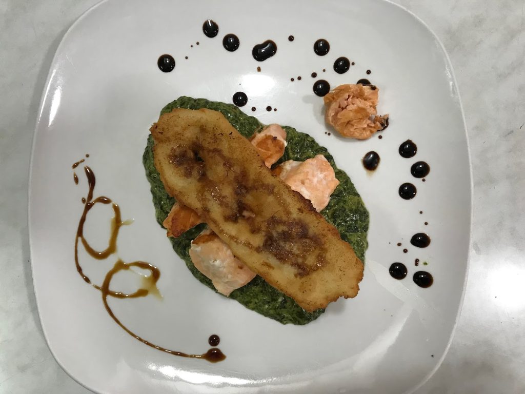 Plato gourmet con bacalao dorado sobre espinacas a la crema y adornos de salsa balsámica.